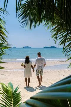 View between palm tree leaves at a couple walking at the beach in Thailand Stock Photos