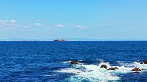 View of a cargo ship sailing across the ocean near rocky shore on a clear day Stock Footage 344806911