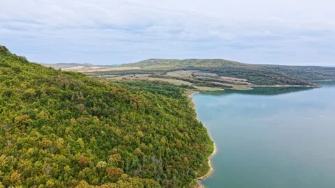 View of a green hillside by a calm lake in a natural setting under a cloudy sky Stock Footage 345497196