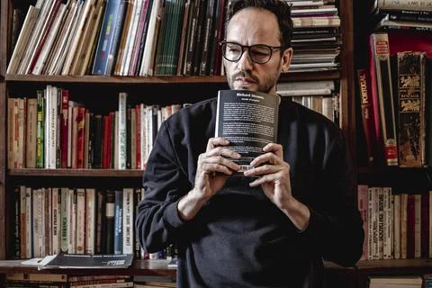 View of an handsome guy reading a book in a library alone wearing glasses, Li Stock Photos