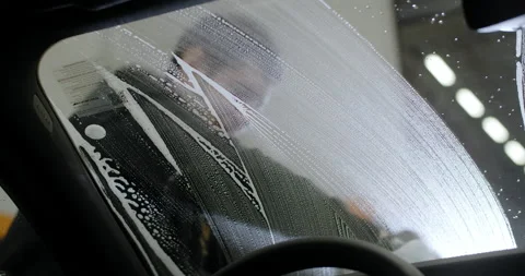 View from inside the car: worker at the carwash cleans vehicle's front window by Stock Footage 172434512