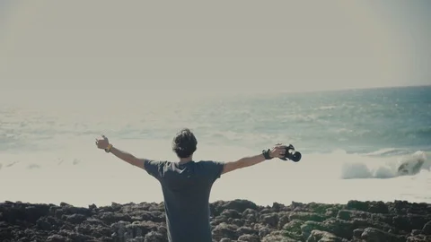 View of the man's back standing in front of the ocean and holding a camera in Stock Footage 90430713