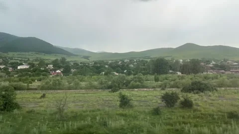 View of mountains, gray sky, field, hill, tree, green grass from train window Video stock 290286070