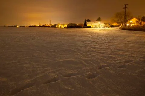 View over a snow covered field with footprints at night to a village with a l Stock Photos