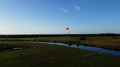 View of a Paraglider Flying at Sunset Over a River and Scenic Green Fields Drone Stock Footage 297624569
