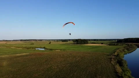 View of a Paraglider Flying at Sunset Over a River and Scenic Green Fields Drone Stock Footage 297624580