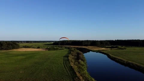 View of a Paraglider Flying at Sunset Over a River and Scenic Green Fields Drone Stock Footage 297624582