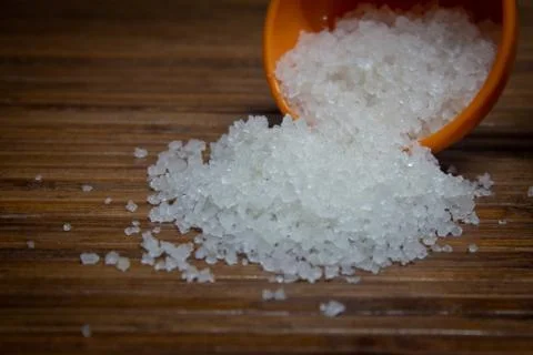 View of the rock salt crystals falling from a measuring cup over a brown wood. Stock Photos