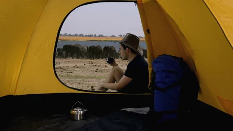 View from tent of guy sitting and looking at elephants nea watering hole, Africa Video stock 161019609