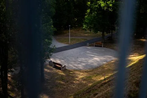 View through a fence of an empty shaded park with alleys and wooden benches Photos