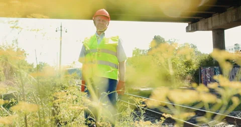 View through grass of senior engineer leans hand and looks at camera on railway Vidéo 135573245