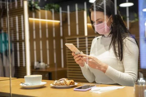 View through shop window of the cafe as a beautiful woman in a medical mask Sits Stock Photos