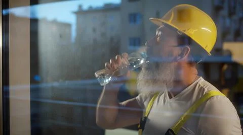View through window of aged construction worker drinking water on location site. Stock Photos