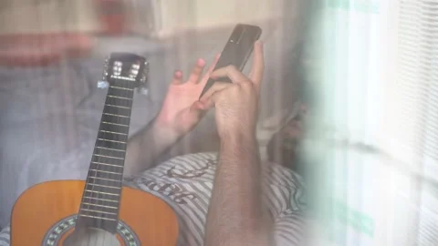 View through a window of a man with guitar lying on sofa using a phone Stock Footage 162169287