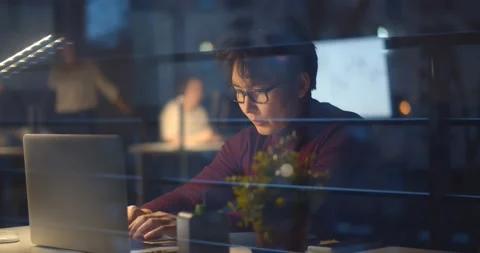 View through window of young asian businessman working late at night in office Stock Footage 147387462