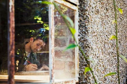 View through a window of a young man drawing in a notebook, a designer at work. Stock Photos