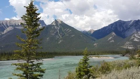View of turquoise river winding through pine forest with rugged mountain peaks. Stock Photos