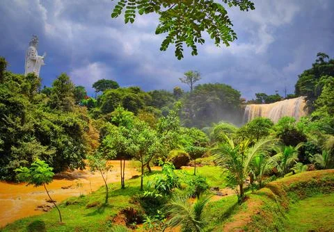 View of a waterfall with a statue surrounded by trees and a river on a clou.. Stock Photos