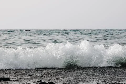 A view of a wave rolling into the ocean on a black volcanic beach. Stock Photos