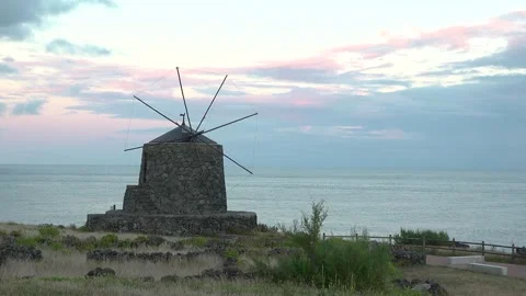 View of windmill near ocean with fields and clouds at sunset on a quiet evening  Stock Footage 335147386