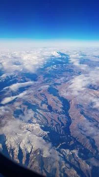 View from window of plane to blue sky and snowy mountain peaks in white clouds Stock Photos