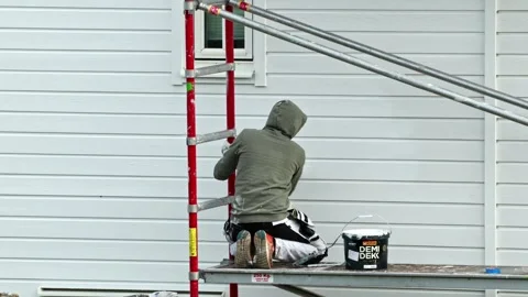 View of worker painting house wall while kneeling on scaffolding platform. Stock Footage 333403850