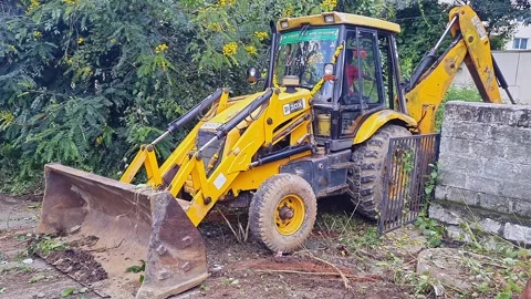 View of Yellow JCB clearing grass near compound wall in outdoor maintenance.. Vídeo Stock 332908699