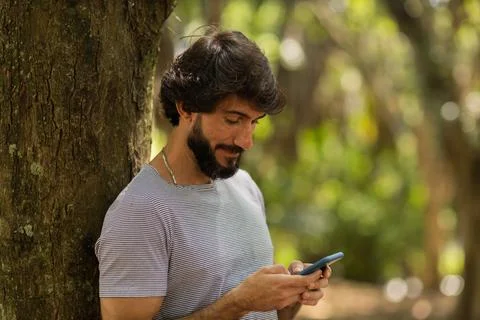 View of young man using a smartphone at day time with a green park in the bac Stock Photos