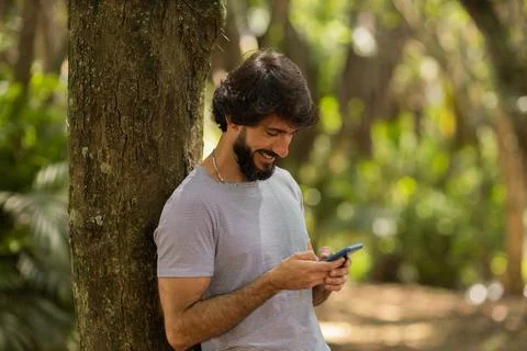View of young man using a smartphone at day time with a green park in the bac Stock Photos