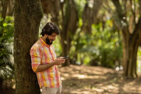 View of young man using a smartphone at day time with a green park in the bac Stock Photos