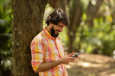 View of young man using a smartphone at day time with a green park in the bac Stock Photos