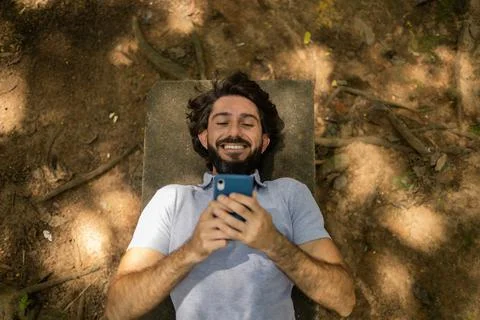 View of young man using a smartphone at day time lying down on a bench at a p Stock Photos