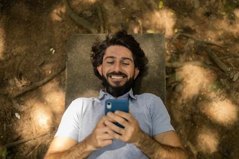View of young man using a smartphone at day time lying down on a bench at a p Stock Photos