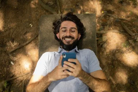 View of young man using a smartphone at day time lying down on a bench at a p Stock Photos