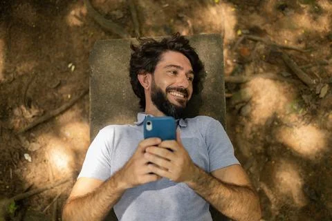 View of young man using a smartphone at day time lying down on a bench at a p Stock Photos