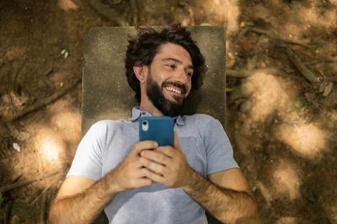 View of young man using a smartphone at day time lying down on a bench at a p Stock Photos