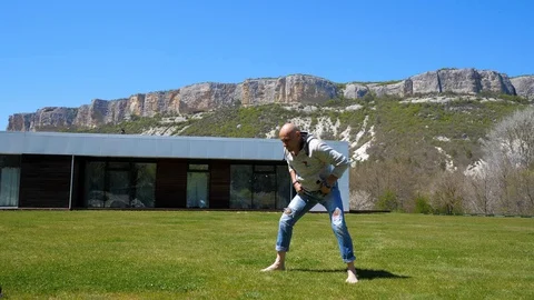 Villa in the mountains. Middle-aged man doing workout on the grass. Capoeirajump Stock Footage 129731565