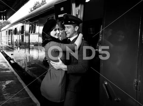 Vintage couple embracing and laughing on railway station platform as train Stock Photos