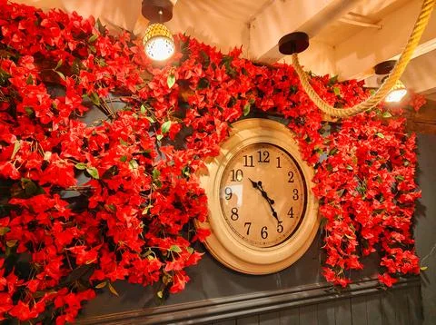 Vintage wall clock surrounded by bright red flowers and warm ceiling lights.. Foto stock
