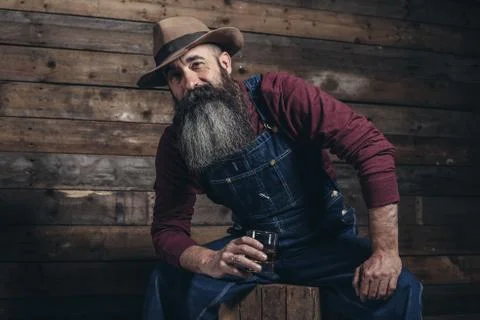 Vintage worker man with long gray beard in jeans dungarees holding whiskey. S Stock Photos