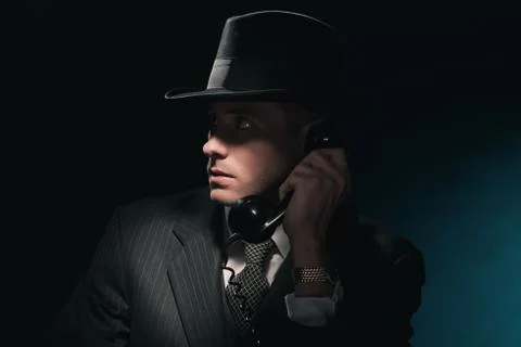 Vintage young detective on the phone with hat in suit and tie. Dark blue back Stock Photos