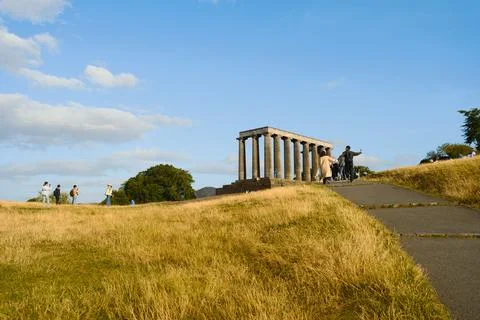 Visitors walking near classical monument on grassy hill under blue sky Stock Photos