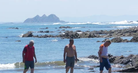 Volcanic Beach And Distant Outcrop Sunny Day As Three Males Cross Through Frame Stock-Footage 138148033