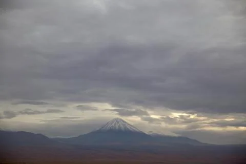 Volcano with snow on the peak under a dramatic sky and sunbeams sneaking Stock Photos
