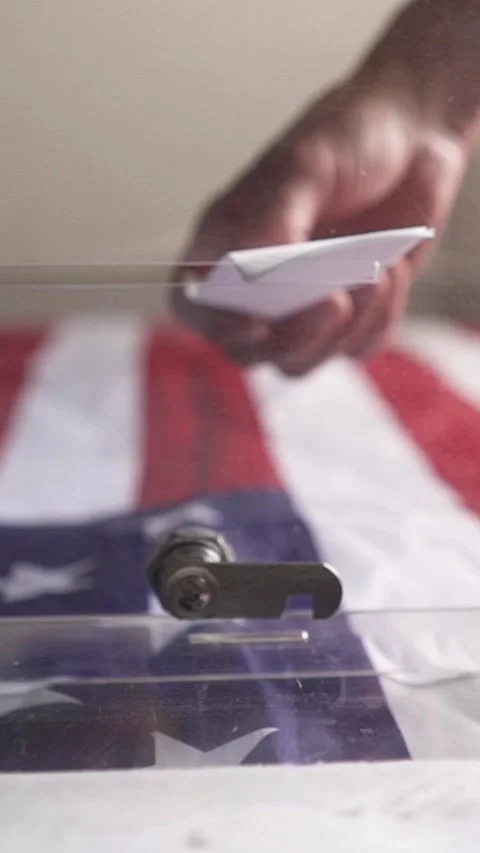 Voter inserting folded ballot paper into transparent box with US flag background Stock Footage 346228209