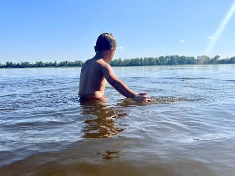 Waist-deep in a river, a boy energetically splashes water around him. Stock Photos