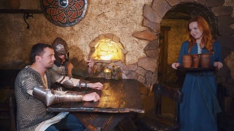 Waitress bringing drinks to table with two medieval warriors in tavern, smiling Stock Footage 79465044