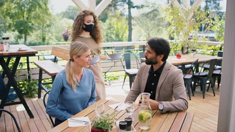 Waitress with face mask serving happy couple outdoors on terrace restaurant. 스톡 동영상 138033507