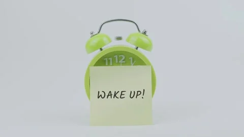 Wake Up! word on sticky note in front of green clock and ringing on white Stock Footage 313919179