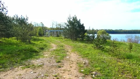 Walking along a dirt path by the water with trees on a sunny day near a wooden s Stock Footage 327181371